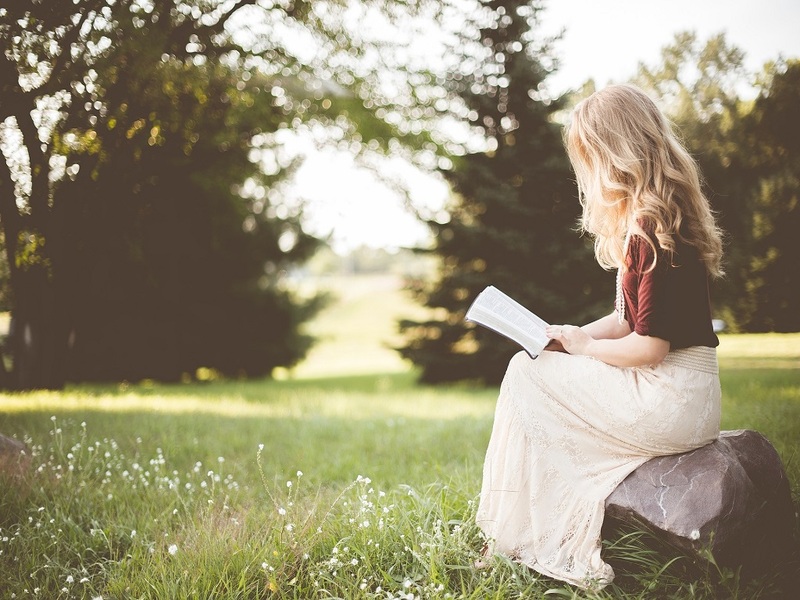 Une jeune femme lit un livre, assise sur une pierre dans une prairie